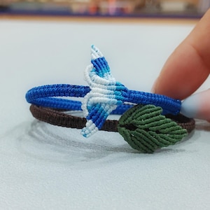 Könnte beinhalten: Ein zweireihiges Makramee-Armband bestehend aus einem blauen Band mit einem weiß-blauen Walflossen-Anhänger und einem braunen Band mit einem grünen Blatt-Anhänger. Die geflochtenen Bänder zeigen durchgehend aufwendige Knüpfmuster.
