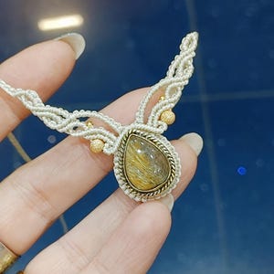 May include: A handmade necklace with a teardrop-shaped pendant, gold-toned frame, and golden-brown stone. It hangs from a cream macrame cord, embellished with small gold beads. This unique piece of jewellery is a statement accessory.