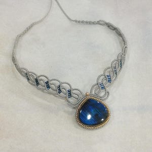 Può includere: Una collana in macramè grigio con un pendente a goccia in pietra preziosa blu. Il pendente è incorniciato con un metallo color oro e un motivo a corda ritorta. La collana presenta perline blu.