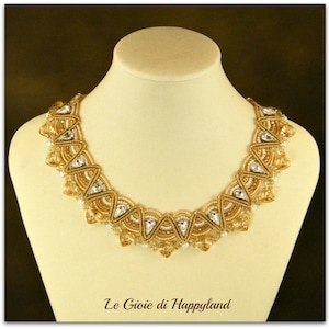 以下が含まれることがあります： 幾何学模様のゴールドとクリアクリスタルのネックレス。ネックレスは白いマネキンバストの上に置かれています。画像の下部に「Le Gioie di Happyland」というテキストがあります。