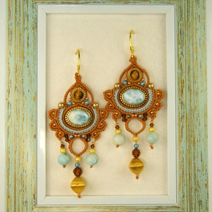 May include: A pair of brown and gold earrings with blue and brown stones hanging from a light blue and white frame.