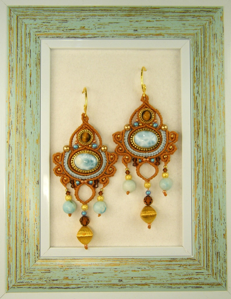 Peut inclure: Une paire de boucles d'oreilles brunes et dor&eacute;es avec des pierres bleues et brunes suspendues &agrave; un cadre bleu clair et blanc.