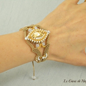 May include: A beige macrame bracelet with a gold and white beaded centerpiece. The bracelet has a tassel and is tied with a knot.
