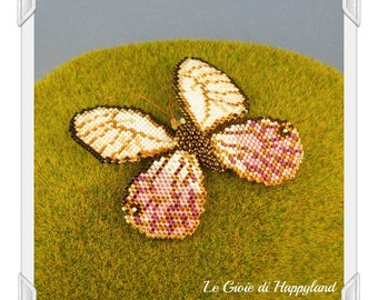 May include: A beaded butterfly brooch with white, gold, and pink accents. The butterfly is lying on a green surface. Le Gioie di Happyland is written in the bottom right corner.