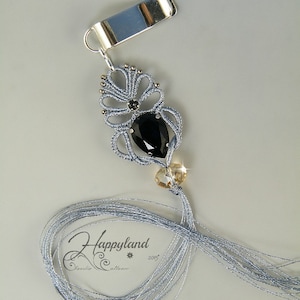 May include: A silver clip with a grey tassel and a black gemstone pendant. The pendant is surrounded by a silver and grey beaded design. The tassel is made of many strands of grey thread. The clip is labelled "Happyland" with a star and the year "2015".