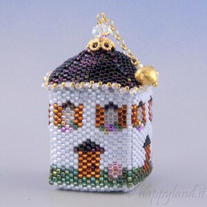 May include: A small, white beaded house ornament with a dark purple roof, gold trim, and a gold chain with a bell. The house has windows with colorful beads and a green lawn.