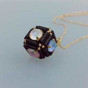May include: A gold chain necklace with a black cube pendant featuring iridescent gemstones. The cube is made of black beads and has a geometric design.