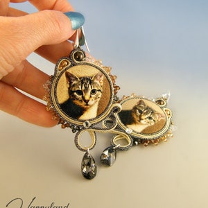 May include: A pair of cat-themed earrings with a gold and brown color scheme. The earrings feature a small, round, framed image of a tabby cat with a black and white pattern. The earrings are accented with small, clear crystals and black beads.