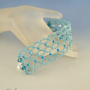 以下が含まれることがあります： 銀色のビーズと青いクリスタルが付いたターコイズブルーのクロシェブレスレット。ブレスレットは白いマネキンの手に置かれています。