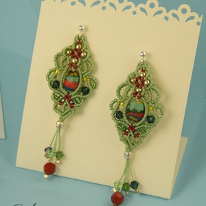 Peut inclure: Boucles d'oreilles en macramé vert et or avec des perles rouges et vertes suspendues au bas. Les boucles d'oreilles sont présentées sur une carte blanche avec le texte "Happyland" et une adresse de site Web.