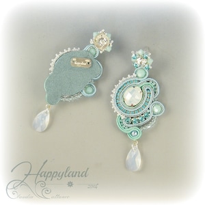 May include: A pair of blue and white earrings with a teardrop-shaped crystal pendant. The earrings are made with a variety of beads and crystals, including turquoise, white, and silver. The earrings are labeled "Happyland 2014".