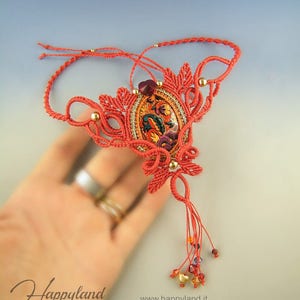 May include: A coral red macrame necklace with a gold and red patterned pendant and a tassel of coral red cord and gold and red beads. The necklace is on a hand with two rings.