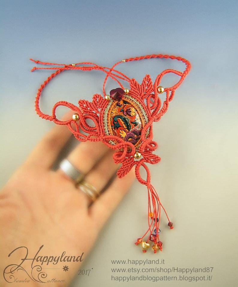May include: A coral red macrame necklace with a gold and red patterned pendant and a tassel of coral red cord and gold and red beads. The necklace is on a hand with two rings.