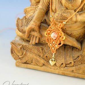 Può includere: Collana in macramé arancione con un ciondolo a forma di cuore e un pendente in pietra rosa. La collana è esposta su una statua di legno.