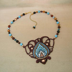 Puede incluir: Un collar artesanal con un colgante central con un diseño floral azul y blanco, enmarcado por macramé marrón. El collar incluye una cadena de cuentas con cuentas turquesas, marrones y negras, y un cierre dorado.