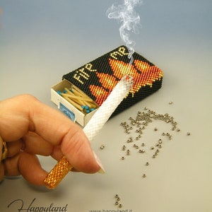 Könnte beinhalten: Eine Perlen-Streichholzschachtel mit dem Wort "FIRE" auf der Vorderseite, eine Perlen-Zigarette und ein Haufen silberner Perlen. Die Streichholzschachtel ist geöffnet und enthält Streichhölzer.