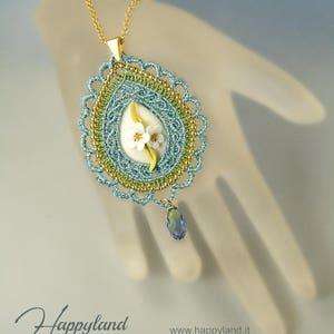 May include: A delicate blue and gold pendant necklace with a white flower design and a blue crystal drop. The pendant is made of crocheted thread and features a floral motif.