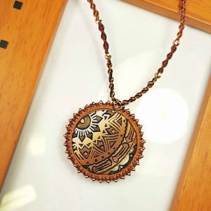 May include: A brown and gold necklace with a round pendant featuring a geometric design. The pendant is made of fabric and has a crocheted border.