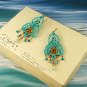 Könnte beinhalten: Ein Paar türkisfarbene Makramee-Ohrringe mit goldfarbenen Akzenten und hängenden Perlen. Die Ohrringe haben einen zentralen bernsteinfarbenen Kristall und werden auf einer cremefarbenen Schachtel mit dem Text "Happyland" und Website-Informationen präsentiert.