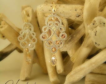 May include: A pair of gold and beige crocheted earrings with crystal accents hanging from a wooden surface. The earrings are delicate and intricate, with a floral design.