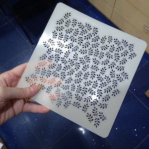 Puede incluir: Una plantilla de plástico blanco con un patrón repetitivo de pequeñas hojas azul oscuro. La plantilla está siendo sostenida por una mano.