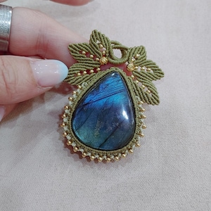 May include: A handmade pendant with a teardrop-shaped labradorite stone, displaying iridescent blue and green colours. The stone is set in a macrame design with olive green cord and gold-coloured beads, and a decorative floral pattern.