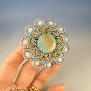 Könnte beinhalten: Eine graue, blaue und goldene Perlenblumenbrosche mit einem grün-gold gestreiften Cabochon in der Mitte. Die Brosche wird in einer Hand gehalten.