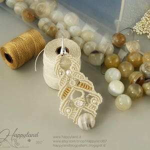 May include: A white and gold macrame earring with a white agate stone pendant. The earring is hanging from a silver hook. The earring is surrounded by white and brown agate beads.