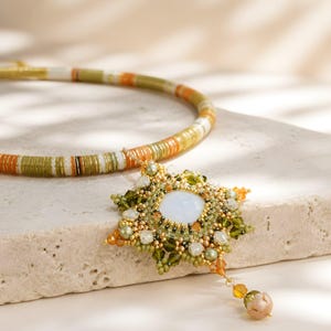 May include: A beaded necklace featuring a circular strand of olive green, orange, and white disc beads. A star-shaped pendant hangs from the strand, centered with a large white cabochon surrounded by intricate green, gold, and orange beadwork.