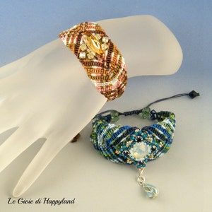 May include: Two handmade friendship bracelets with intricate knot patterns and colorful beads. The top bracelet features a brown, pink, and white pattern with a large, clear crystal. The bottom bracelet has a blue, green, and gold pattern with a smaller, clear crystal pendant.