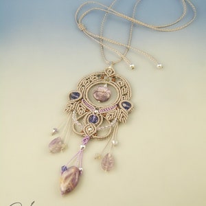 Op de afbeelding: Een beige macramé ketting met een grote, ingewikkelde hanger met een paarse en bruine steen. De hanger is versierd met kleinere paarse en heldere stenen en hangt aan een beige koord.