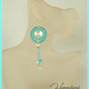May include: Teal and green beaded earrings with a crystal pendant and a silver hook. The earrings are hanging from a mannequin's ear.