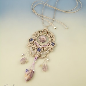 May include: A beige macrame necklace with a large, intricate pendant featuring a purple and brown stone. The pendant is accented with smaller purple and brown stones and silver beads. The necklace is tied with a simple knot at the top.