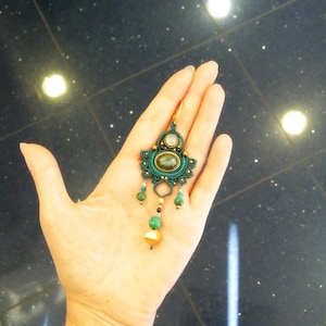 Peut inclure: Une boucle d'oreille &agrave; perles vertes et dor&eacute;es avec un grand pendentif en forme de pierre ovale. La boucle d'oreille est suspendue &agrave; une main.