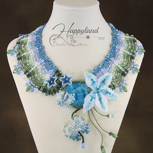 May include: A blue and green beaded necklace with a large blue stone pendant and a white flower design. The necklace is on a white mannequin bust. The text "Happyland Claudia Attance 2014" is on the necklace.
