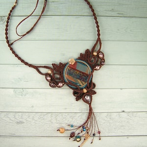 May include: A brown macrame necklace with a large, round, multi-colored stone pendant. The pendant has a blue, green, and brown pattern. The necklace has a tassel of brown cords and beads.