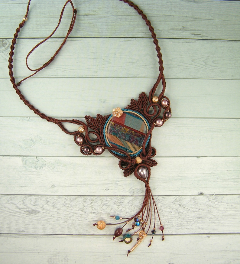 May include: A brown macrame necklace with a large, round, multi-colored stone pendant. The pendant has a blue, green, and brown pattern. The necklace has a tassel of brown cords and beads.