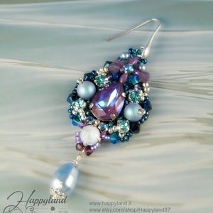 May include: A single drop earring featuring a central pear-shaped purple crystal surrounded by light blue pearls, dark blue beads, and smaller turquoise crystals. A larger light blue pearl hangs from the bottom. Includes text: Happyland 2018.