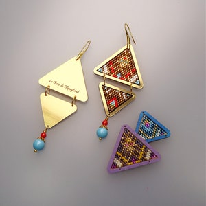 May include: A pair of gold triangle earrings with colourful beaded designs. The earrings feature a smaller triangle with a beaded design on top of a larger triangle with a gold metal surface. The larger triangle has the text "Le Gioie di Happyland" engraved on it.