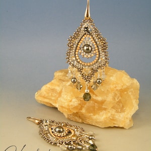 Könnte beinhalten: Ein handgefertigtes Perlen-Anhänger-Halsketten- und Ohrring-Set. Der Anhänger hat eine Tropfenform mit einer zentralen dunkelgrauen Perle, umgeben von goldenen, silbernen und pfirsichfarbenen Perlen. Die Ohrringe spiegeln das Design des Anhängers wider. Das Set wird auf einem hellen Stein präsentiert.