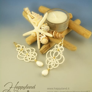 May include: A pair of white, lace-like earrings with gold accents and a teardrop-shaped pearl. The earrings are displayed with seashells, a starfish, a candle, and driftwood, creating a beachy theme.