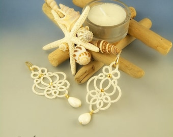 May include: A pair of white, lace-like earrings with gold accents and a teardrop-shaped pearl. The earrings are displayed with seashells, a starfish, a candle, and driftwood, creating a beachy theme.