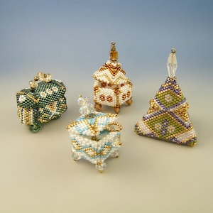Op de afbeelding: Vier kleine kralen doosjes in verschillende vormen en kleuren. De doosjes zijn gemaakt van witte, groene, gouden en paarse kralen. De doosjes zijn allemaal gesloten en hebben een klein lusje bovenop.