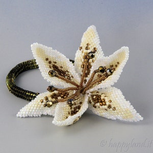 Könnte beinhalten: Eine weiße Perlenlilie mit goldenen Akzenten, befestigt an einem grünen Gummiband. Die Blume ist aus kleinen, schillernden Perlen gefertigt und hat einen zarten, femininen Look.