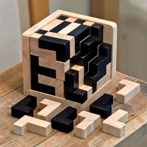 May include: A wooden cube puzzle with black and natural wood-colored pieces. The puzzle is partially assembled, with some pieces removed and scattered around. The cube is sitting on a wooden surface.