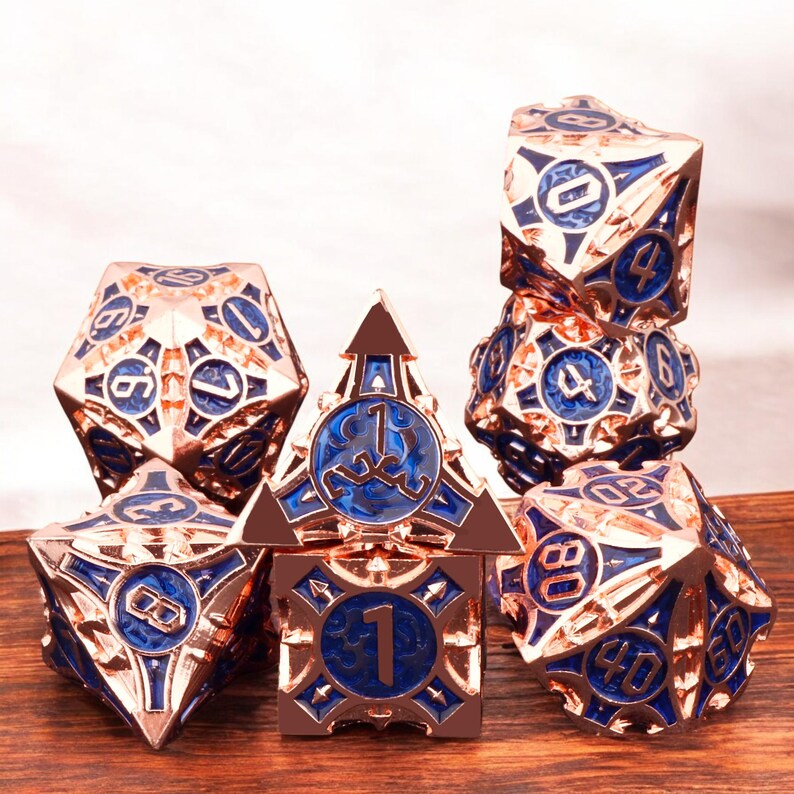 May include: Set of seven rose gold and blue metal dice with intricate designs. The dice are numbered 1 through 6 and include a d4, d6, d8, d10, d%, d12, and d20.