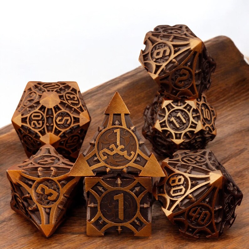 May include: A set of seven metal dice with a bronze finish. The dice are detailed with intricate designs and have a raised number on each side. The dice are arranged on a wooden surface.