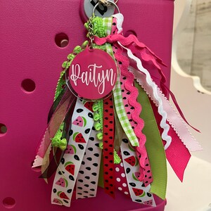 May include: A pink and green personalized keychain with the name "Paityn" on a round acrylic tag. The keychain features a variety of ribbons with watermelon prints, polka dots, and gingham patterns.