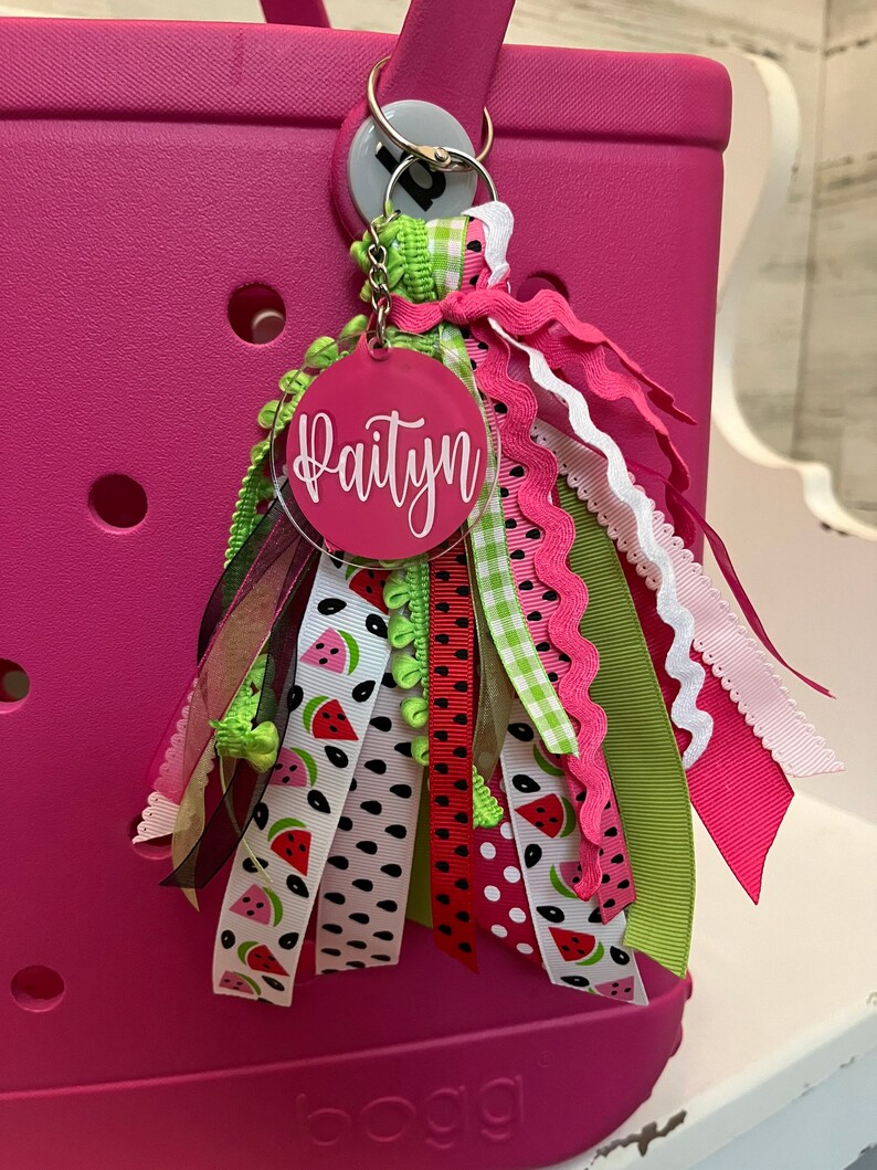 May include: A pink and green personalized keychain with the name "Paityn" on a round acrylic tag. The keychain features a variety of ribbons with watermelon prints, polka dots, and gingham patterns.