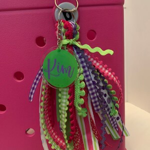 Pode incluir: Uma bolsa tote rosa com um chaveiro decorativo. O chaveiro apresenta uma etiqueta circular verde com o nome "Kim" em escrita cursiva e fitas coloridas em tons de rosa, roxo, verde e branco. A bolsa tem uma al&ccedil;a e um anel prateado.